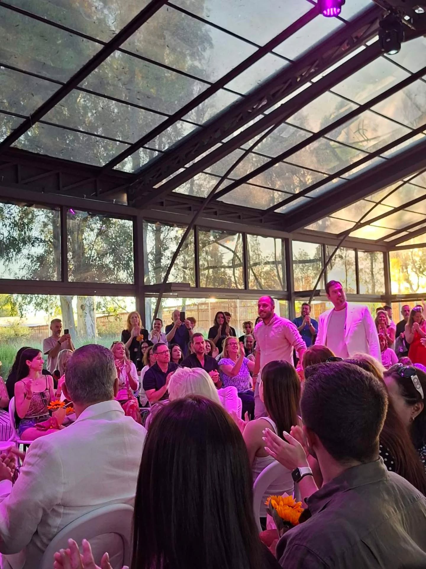 Crowd gathered in a glass-walled venue, with two men in light suits standing while guests applaud.