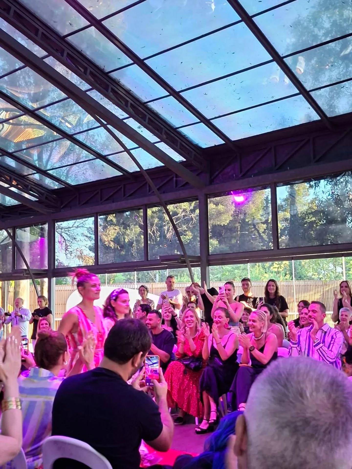 Audience watching a fashion show under a glass ceiling, with a model walking the runway and pink stage lighting visible.