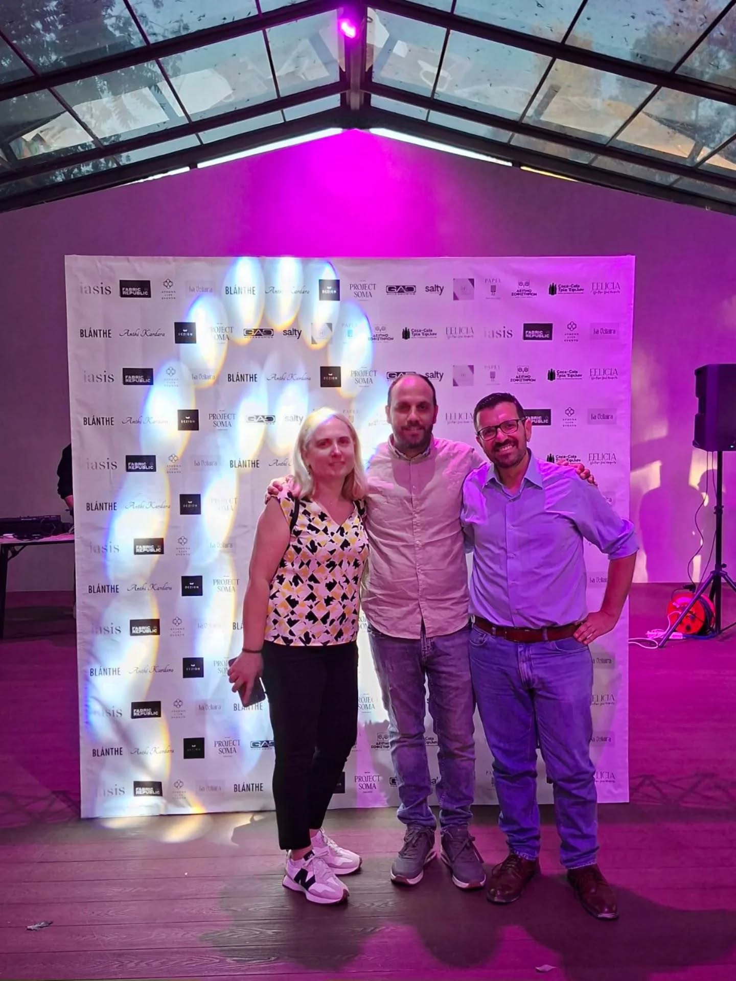 Three people posing together in front of a sponsor banner at a purple-lit event.