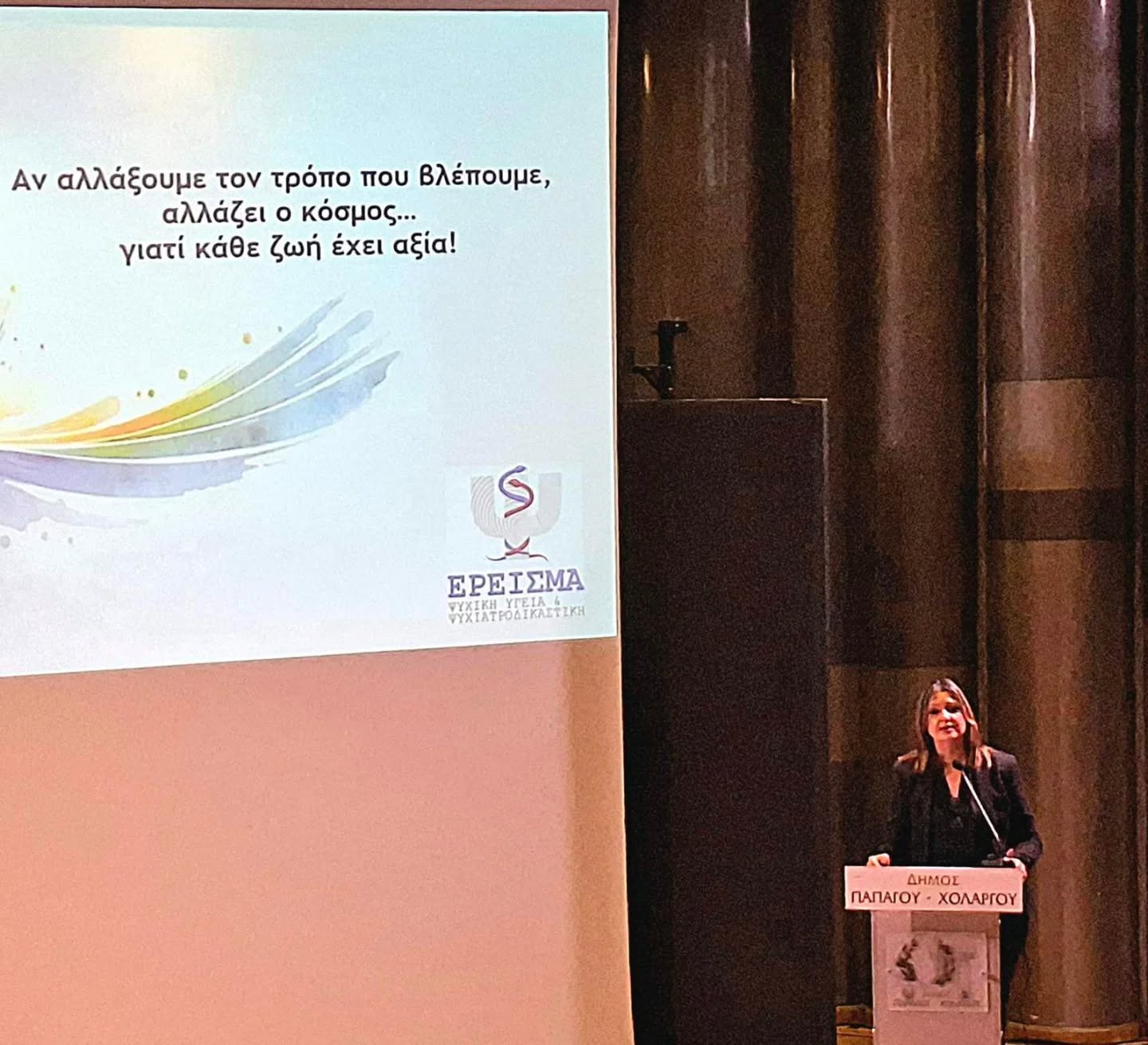 Female presenter stands at a podium on a dimly lit stage, delivering a talk; a slide with Greek text and colorful swooshes is projected on a screen to the left.