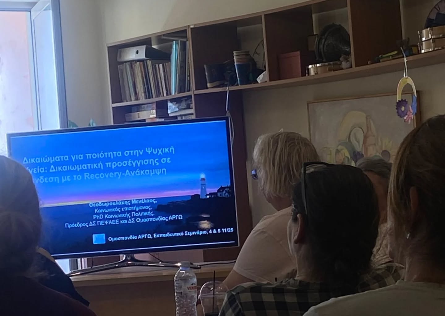People sit and watch a Greek-language presentation on a flat-screen TV in a living-room setup with shelves in the background.