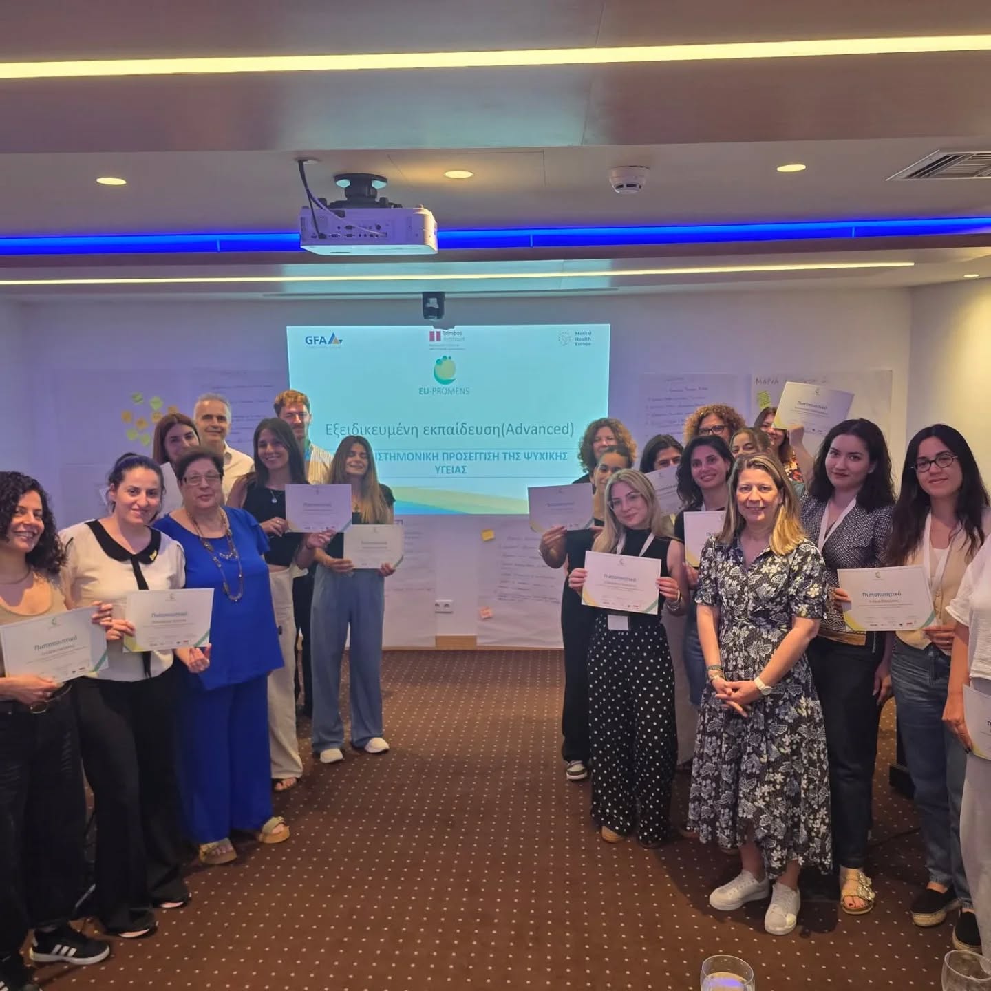 Group of diverse adults in a conference room posing with certificates after a training program; projector screen with logos in the back.