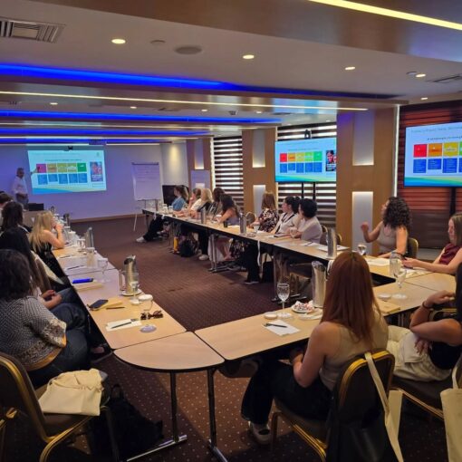 Conference room with attendees seated around a U-shaped table listening to a presenter; colorful charts displayed on screens at the front and side of the room.