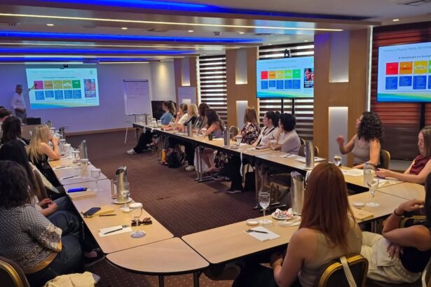 Conference room with attendees seated around a U-shaped table listening to a presenter; colorful charts displayed on screens at the front and side of the room.