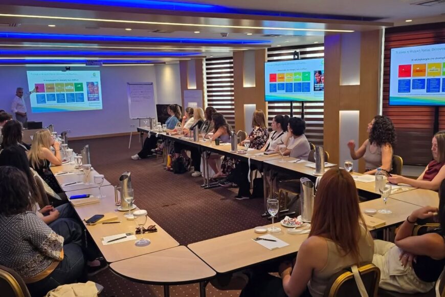Conference room with attendees seated around a U-shaped table listening to a presenter; colorful charts displayed on screens at the front and side of the room.