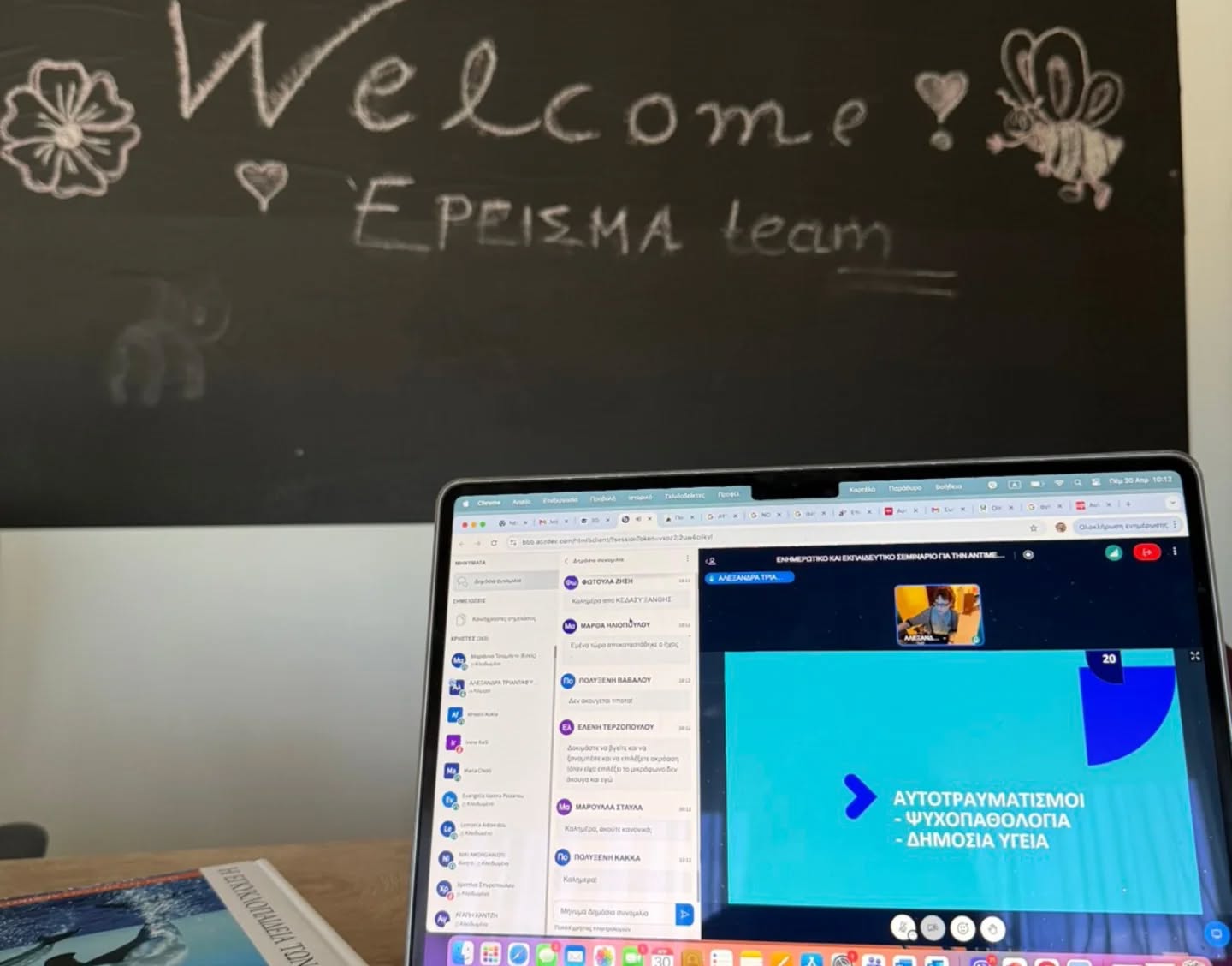 Chalkboard with a 'Welcome' message and doodles, in front of a laptop open on a desk showing a Greek presentation slide.