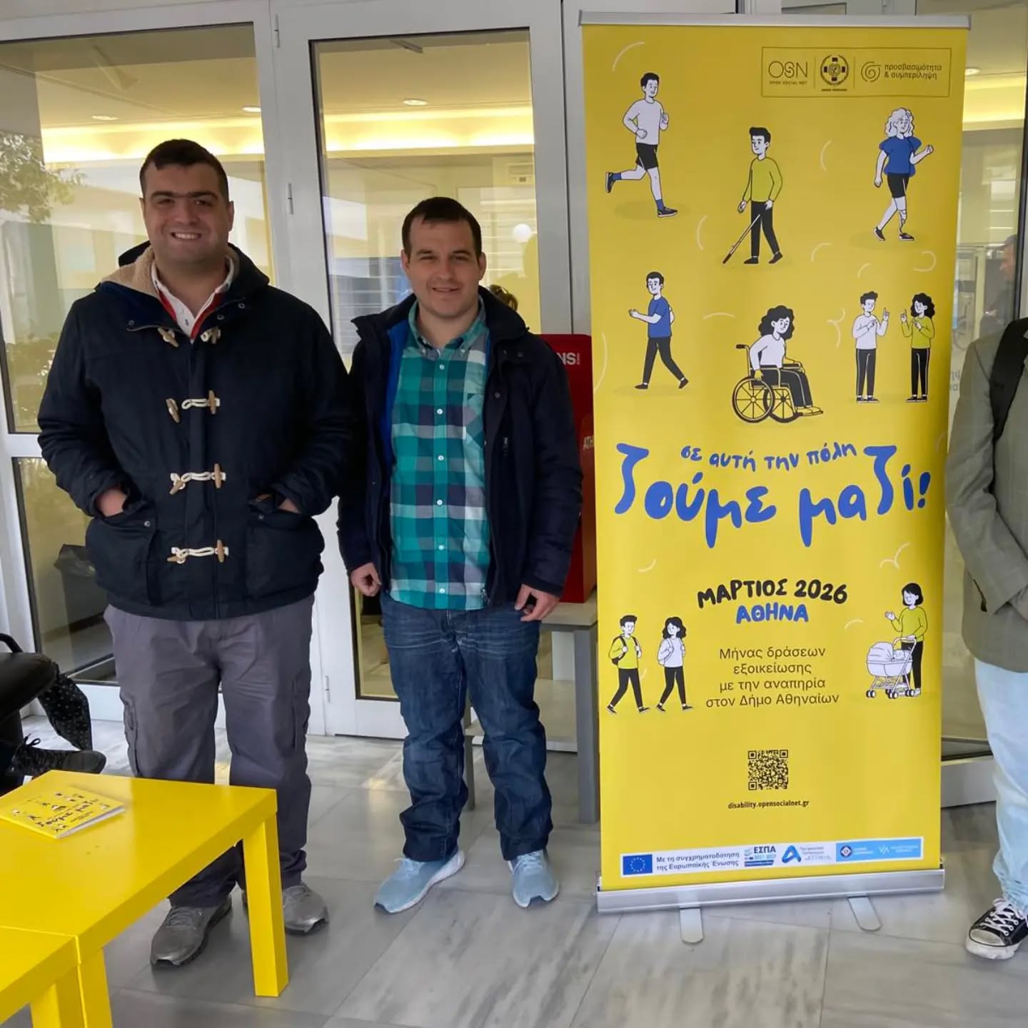 Two men pose indoors beside a bright yellow disability-awareness banner in Athens (March 2026).