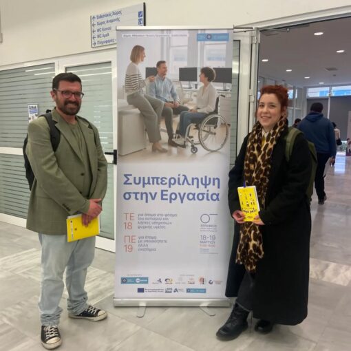 Man in a green blazer and jeans and a woman in a black coat with a leopard print scarf stand beside a tall banner about inclusion in the workplace, each holding a yellow brochure.