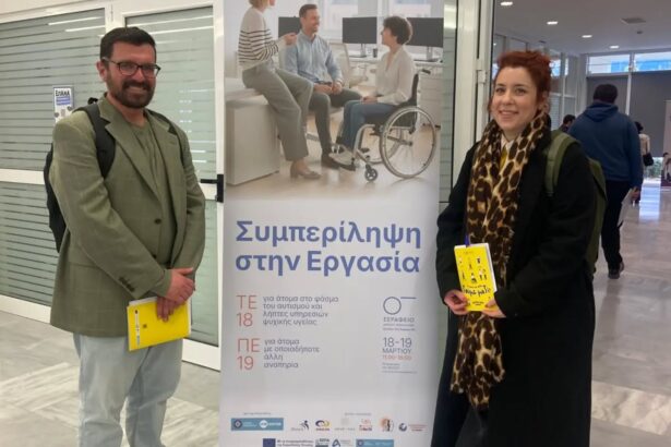 Man in a green blazer and jeans and a woman in a black coat with a leopard print scarf stand beside a tall banner about inclusion in the workplace, each holding a yellow brochure.