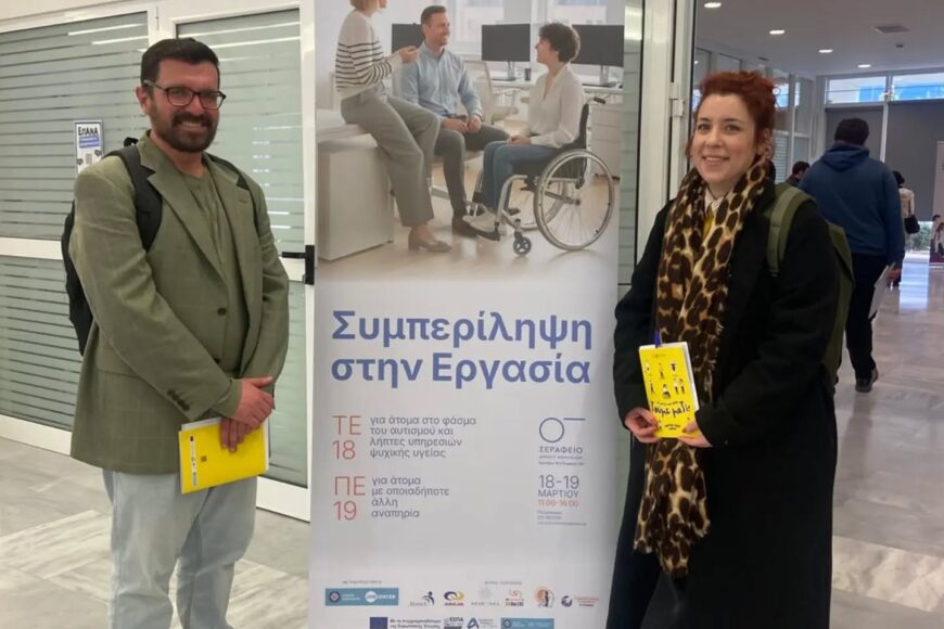 Man in a green blazer and jeans and a woman in a black coat with a leopard print scarf stand beside a tall banner about inclusion in the workplace, each holding a yellow brochure.