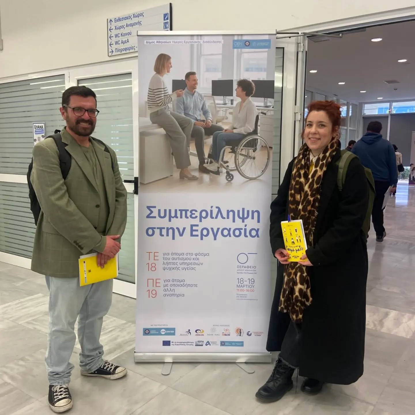 Man in a green blazer and jeans and a woman in a black coat with a leopard print scarf stand beside a tall banner about inclusion in the workplace, each holding a yellow brochure.