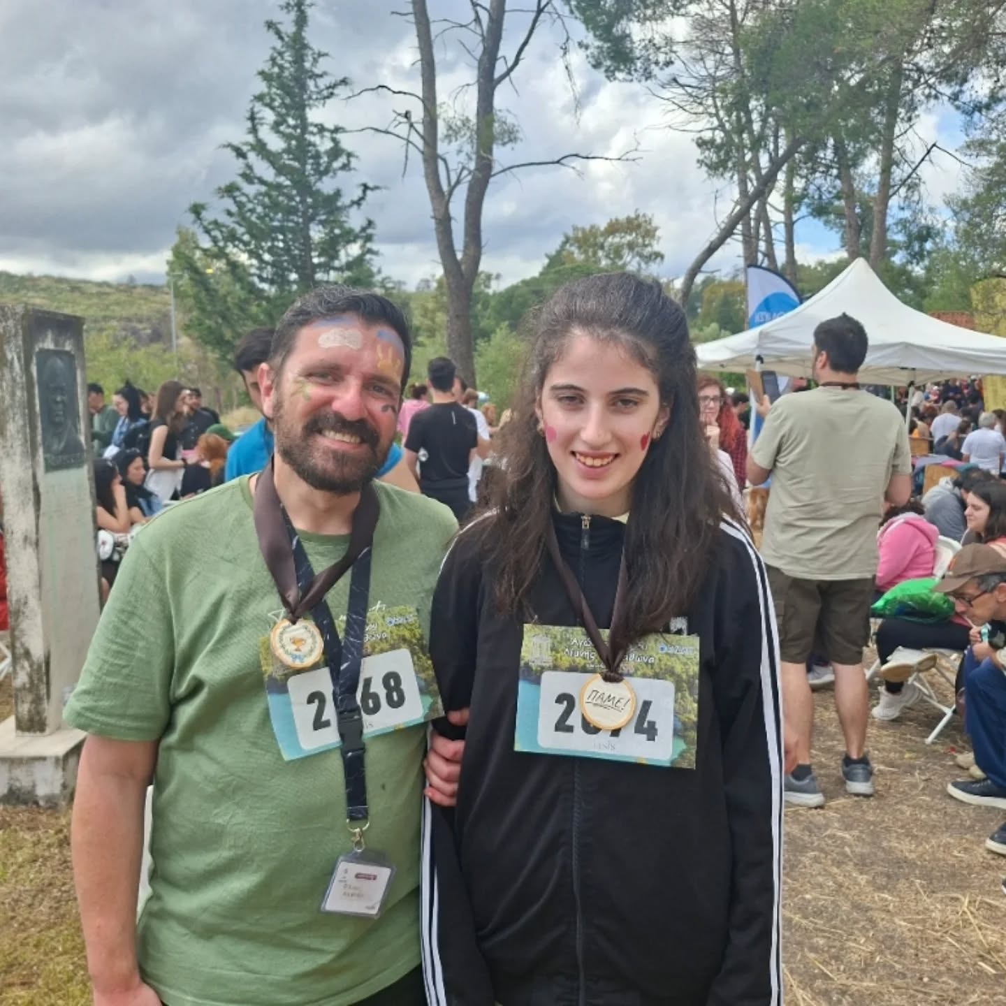Two smiling participants at an outdoor festival wearing race bibs and medals, face paint on cheeks, posing together.