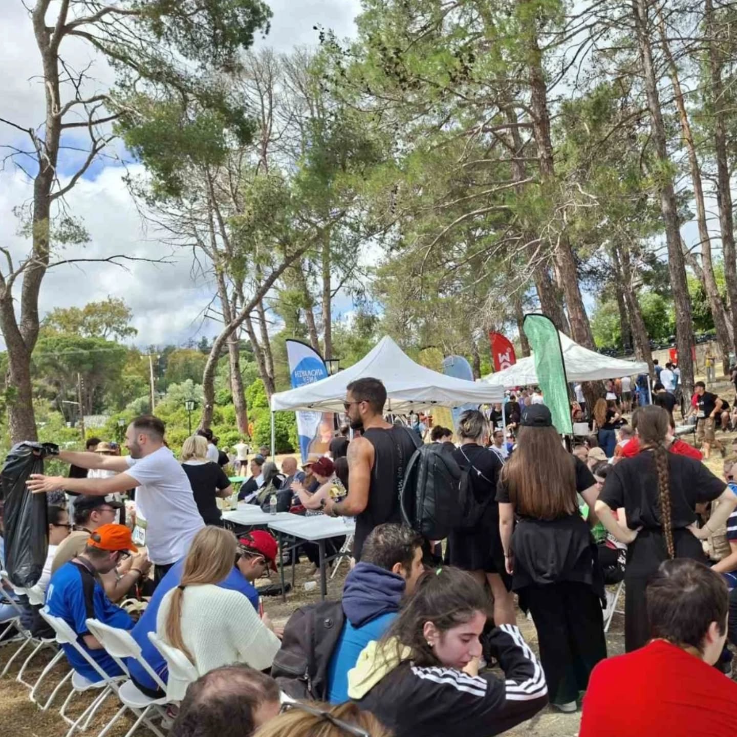 Crowd gathered at an outdoor festival with white tents, folding chairs, and tall trees in a park-like setting.