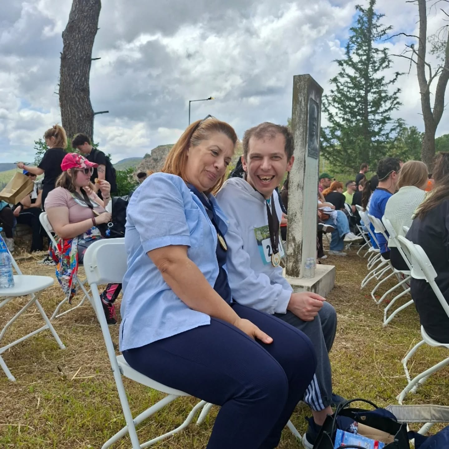 Two people in the foreground smile at the camera while seated on white folding chairs at an outdoor event, with more attendees behind them.