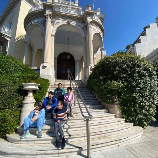 Five people sit on curved marble steps of a historic building with tall decorative columns and an arched entryway, greenery on both sides.