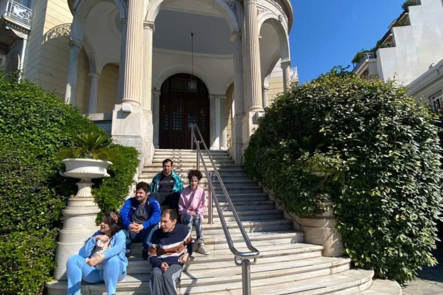 Five people sit on curved marble steps of a historic building with tall decorative columns and an arched entryway, greenery on both sides.