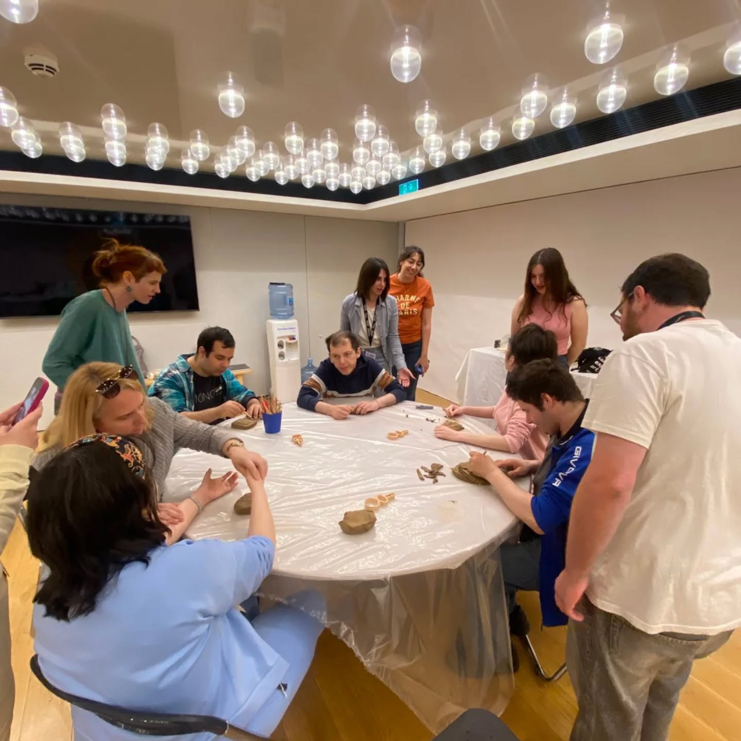 Group of people around a plastic-covered round table, shaping clay in a workshop room with hanging bulb lights.