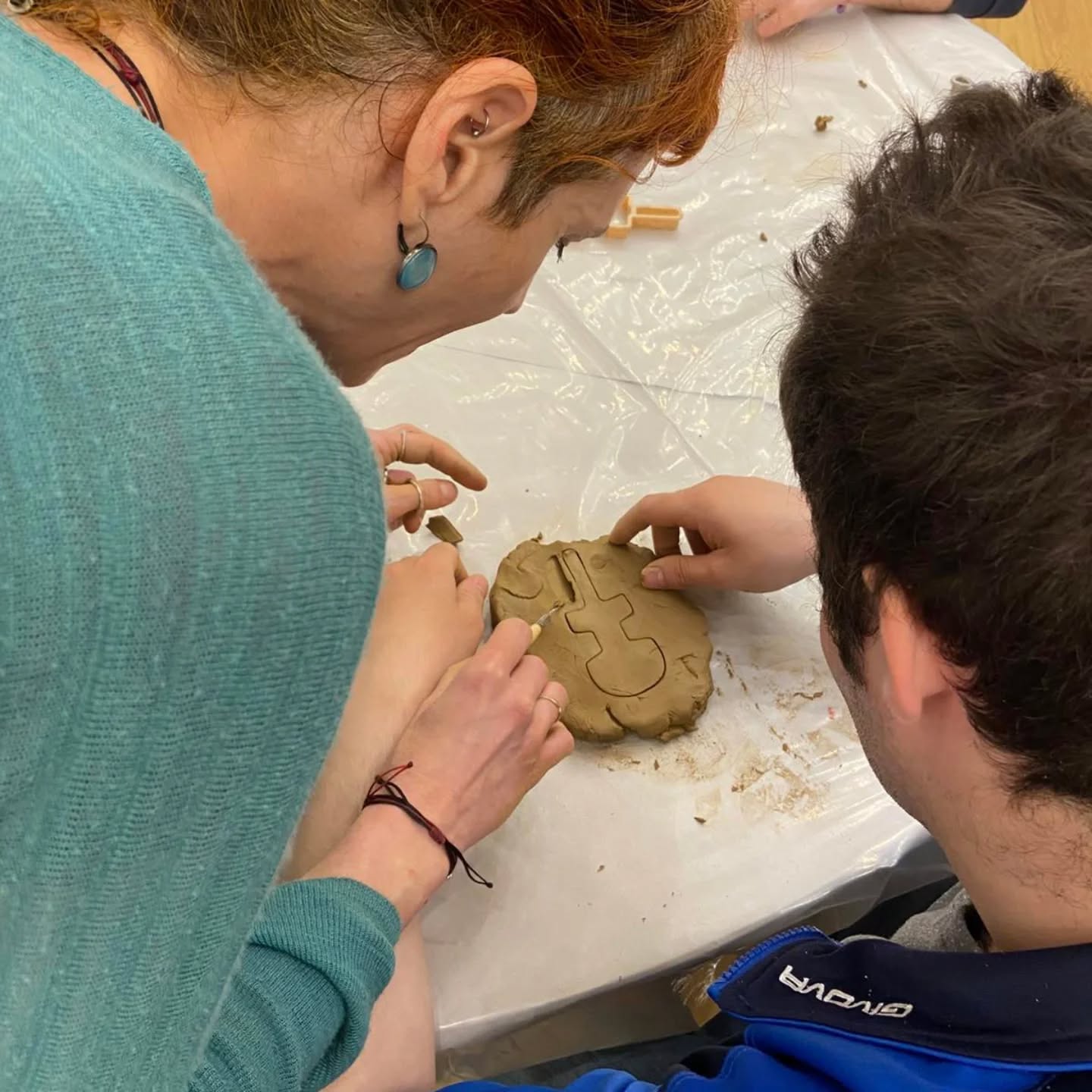 Two people sculpting a round clay piece with tools on a plastic-covered table, focusing on the details of the carving work.