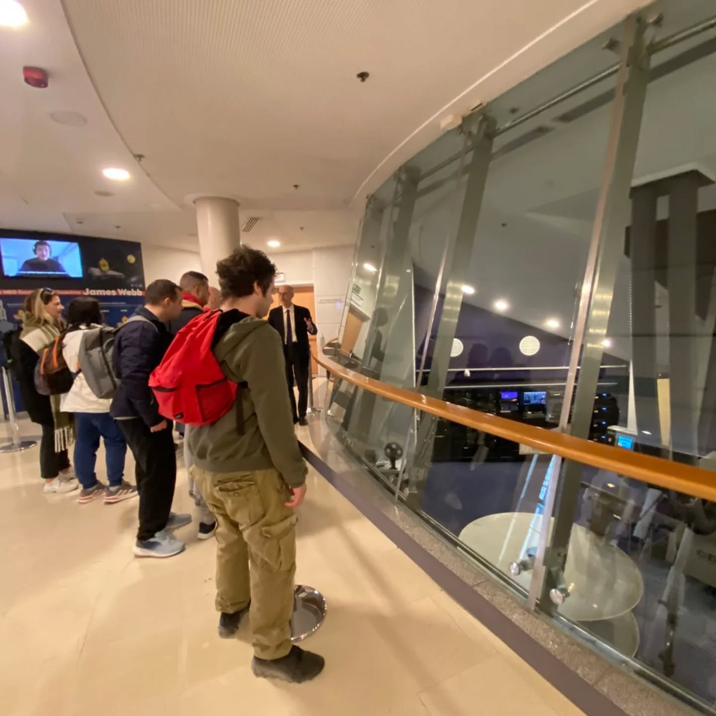 Group of travelers with backpacks standing in a queue beside a curved glass barrier in an airport or terminal.