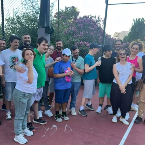 Group of people posing together on an outdoor basketball court, smiling with a chain-link fence and pink flowering bushes in the background.