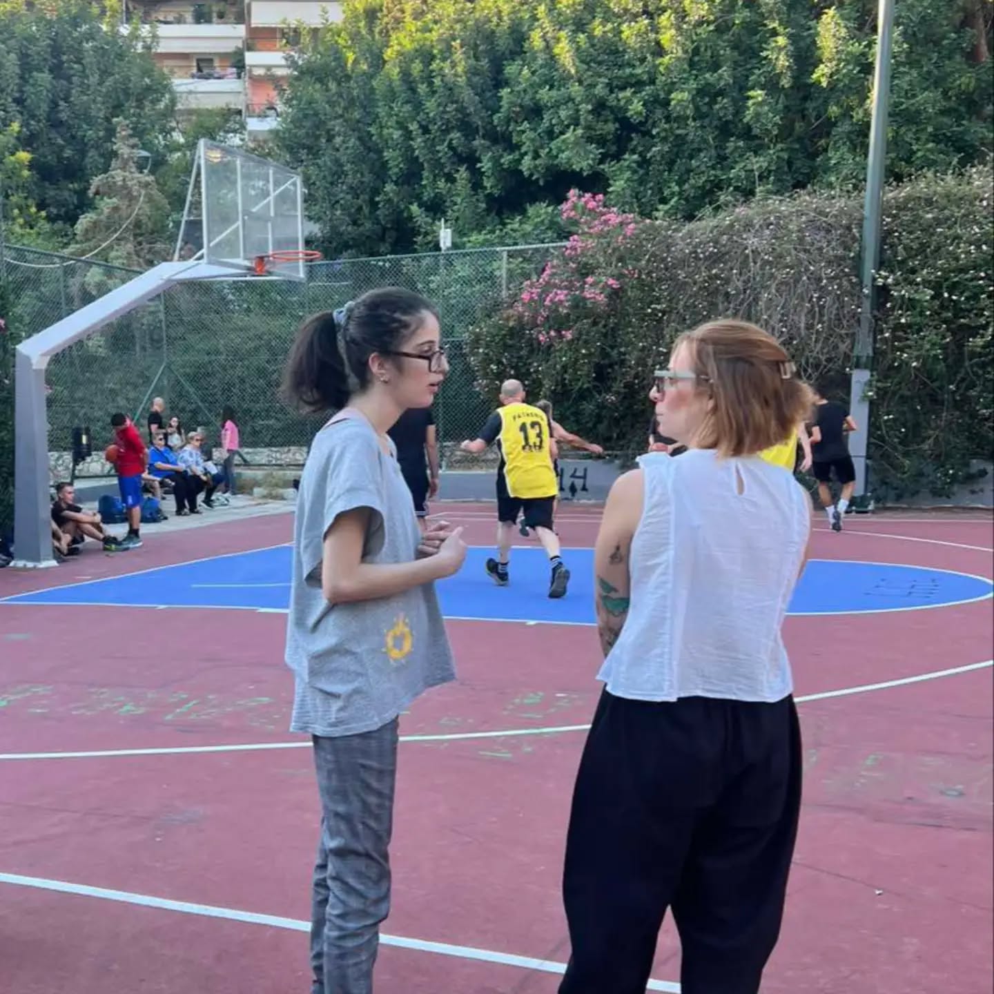 Two women chat on an outdoor basketball court as players run in the background, with a hoop and red surface nearby.