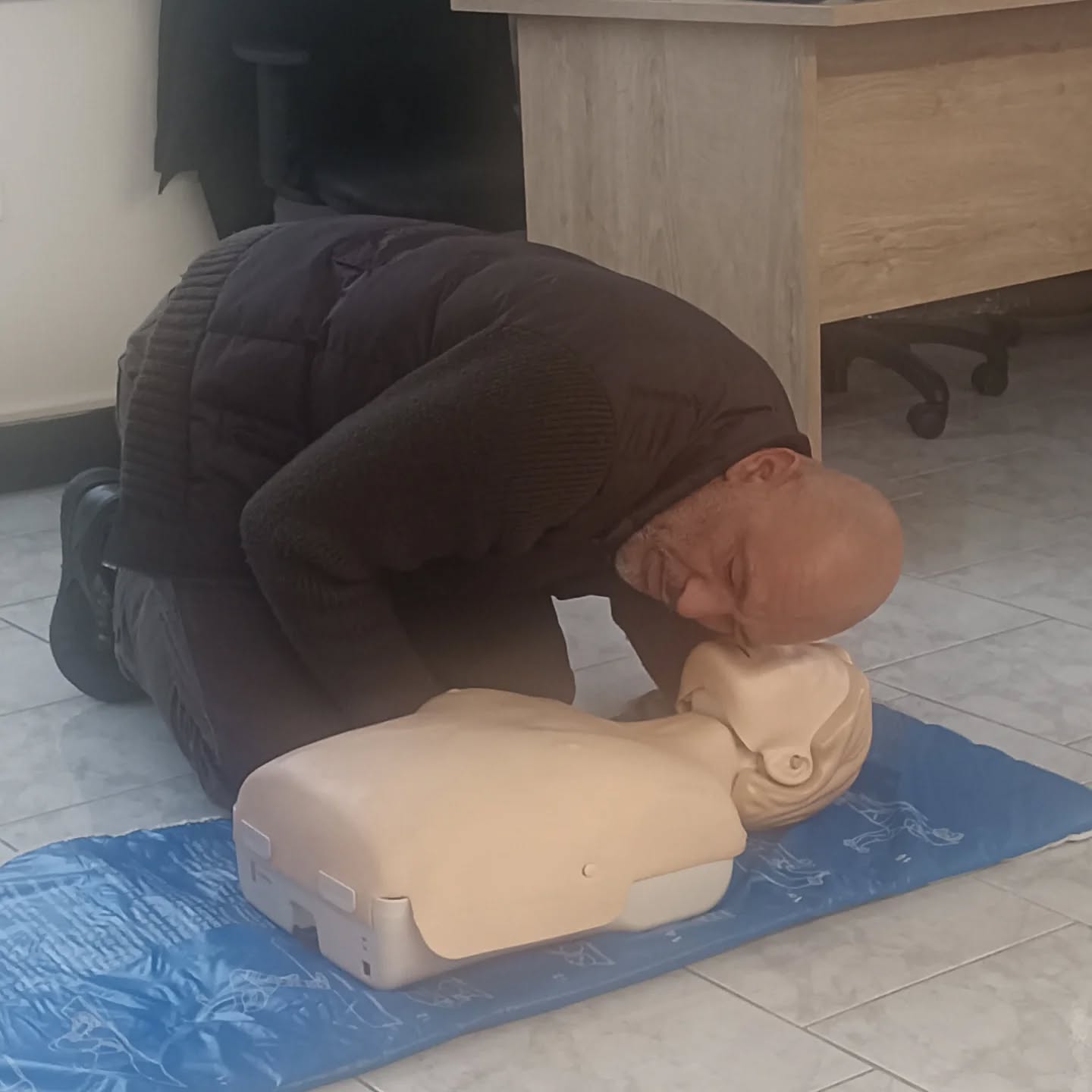 Man kneels on a blue mat, giving rescue breaths to a CPR mannequin during training session.