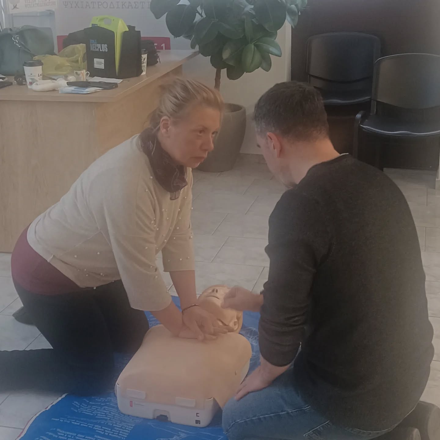 Two adults kneel over a CPR manikin, practicing chest compressions on a blue mat in a training room.