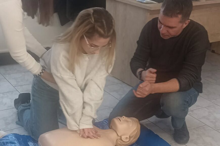Two people kneeling on the floor performing chest compressions on a CPR manikin during a first-aid training session.