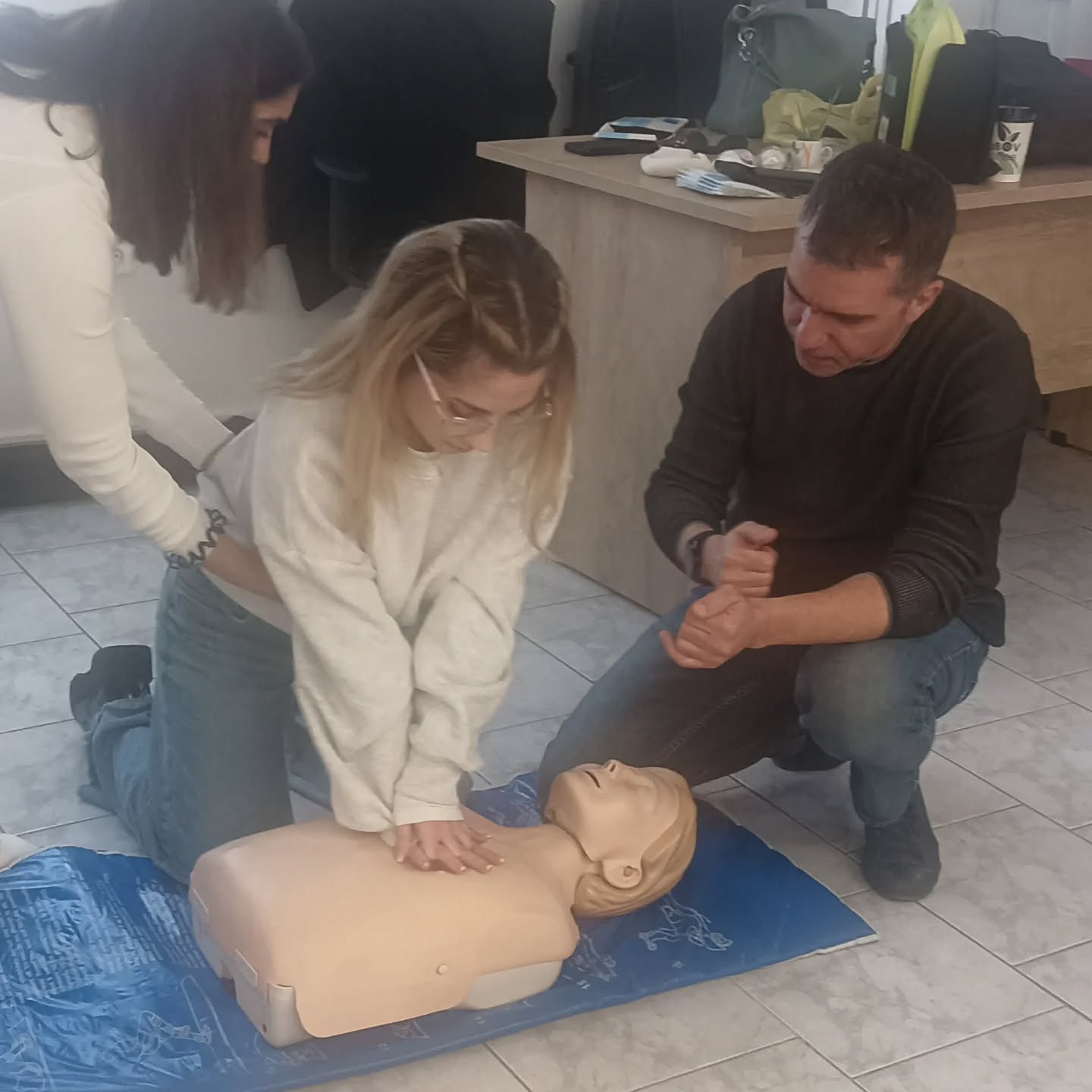 Two people kneeling on the floor performing chest compressions on a CPR manikin during a first-aid training session.