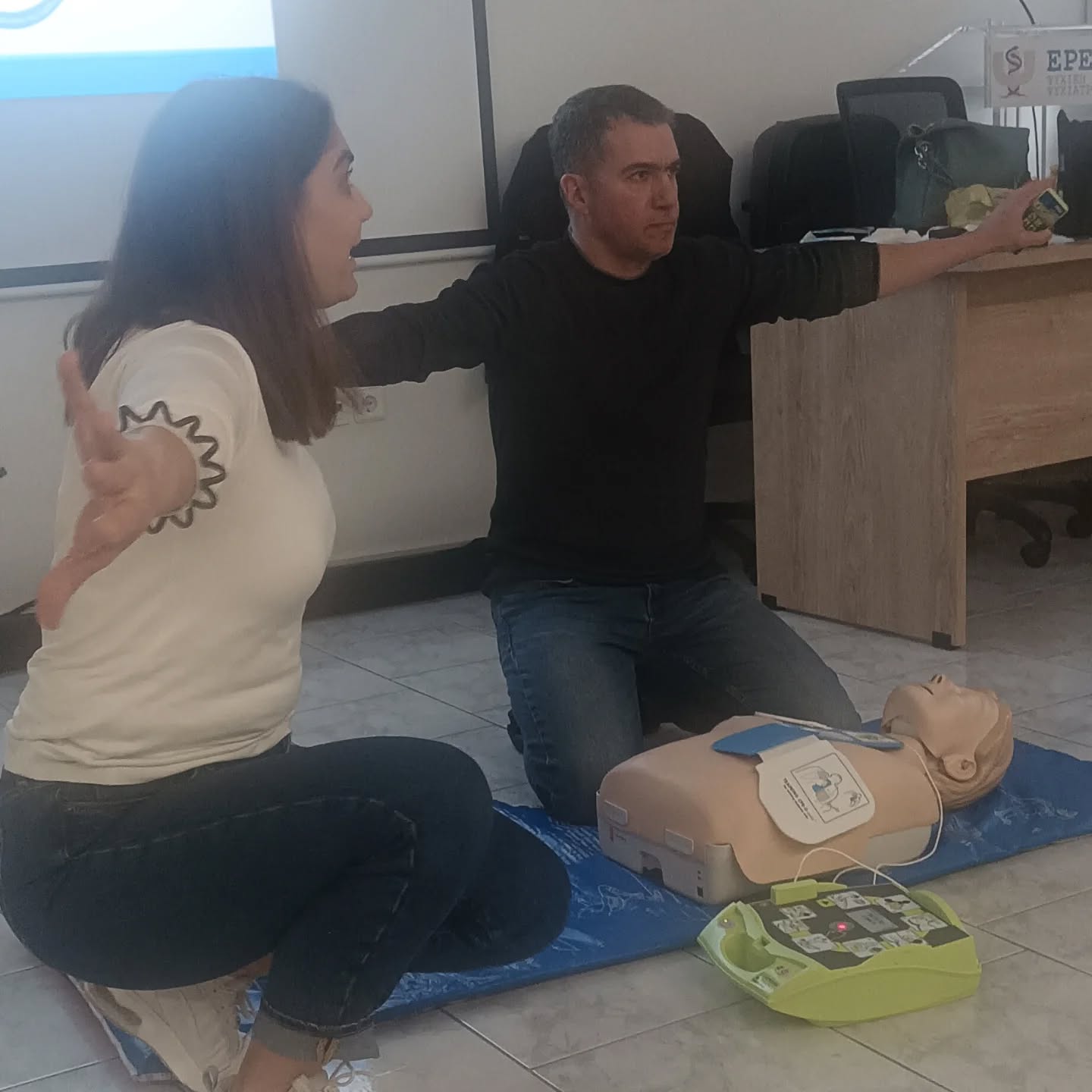 Two adults kneeling on the floor, practicing CPR on a manikin with an AED trainer nearby.