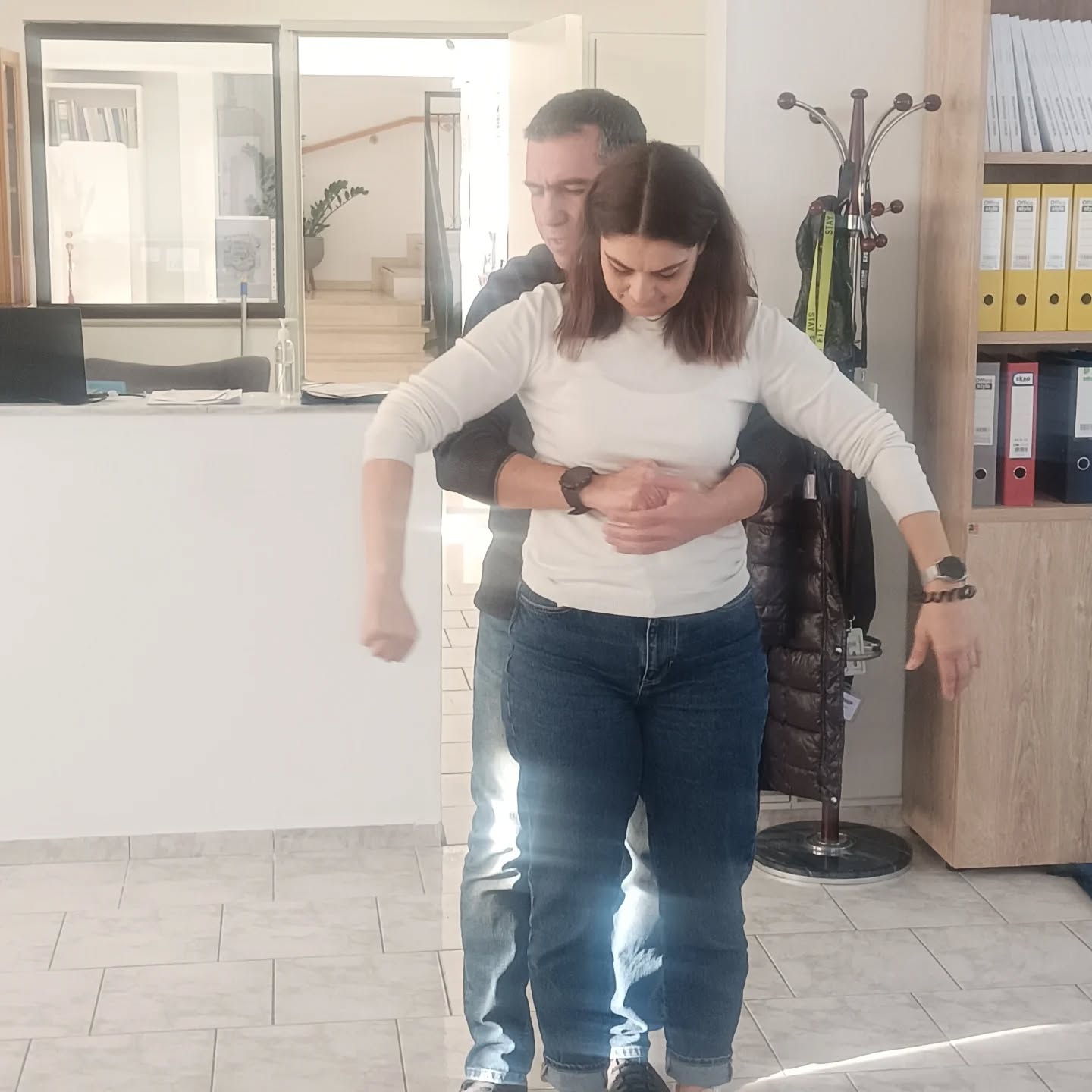 A man stands behind a woman, guiding her by holding her around the waist in a bright office reception area with shelves and a coat rack nearby.
