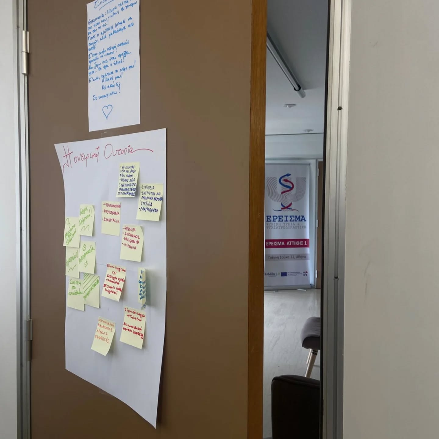 Doorway with a large white poster covered in yellow sticky notes on a brown door; a handwritten sheet above. In the background, a banner with Greek text and a logo is visible.