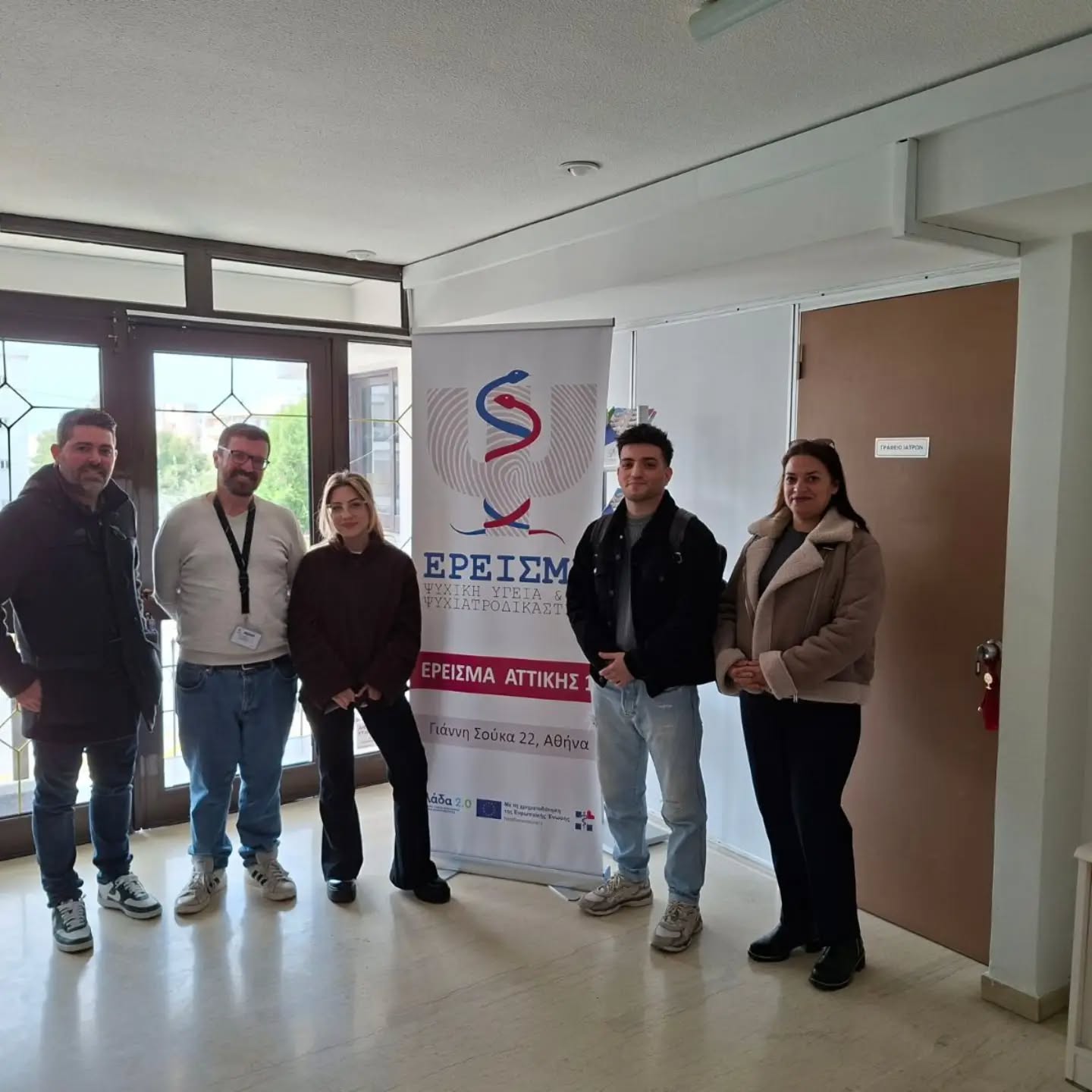 Five people stand indoors beside a tall banner with a logo, posing for a group photo at an event.
