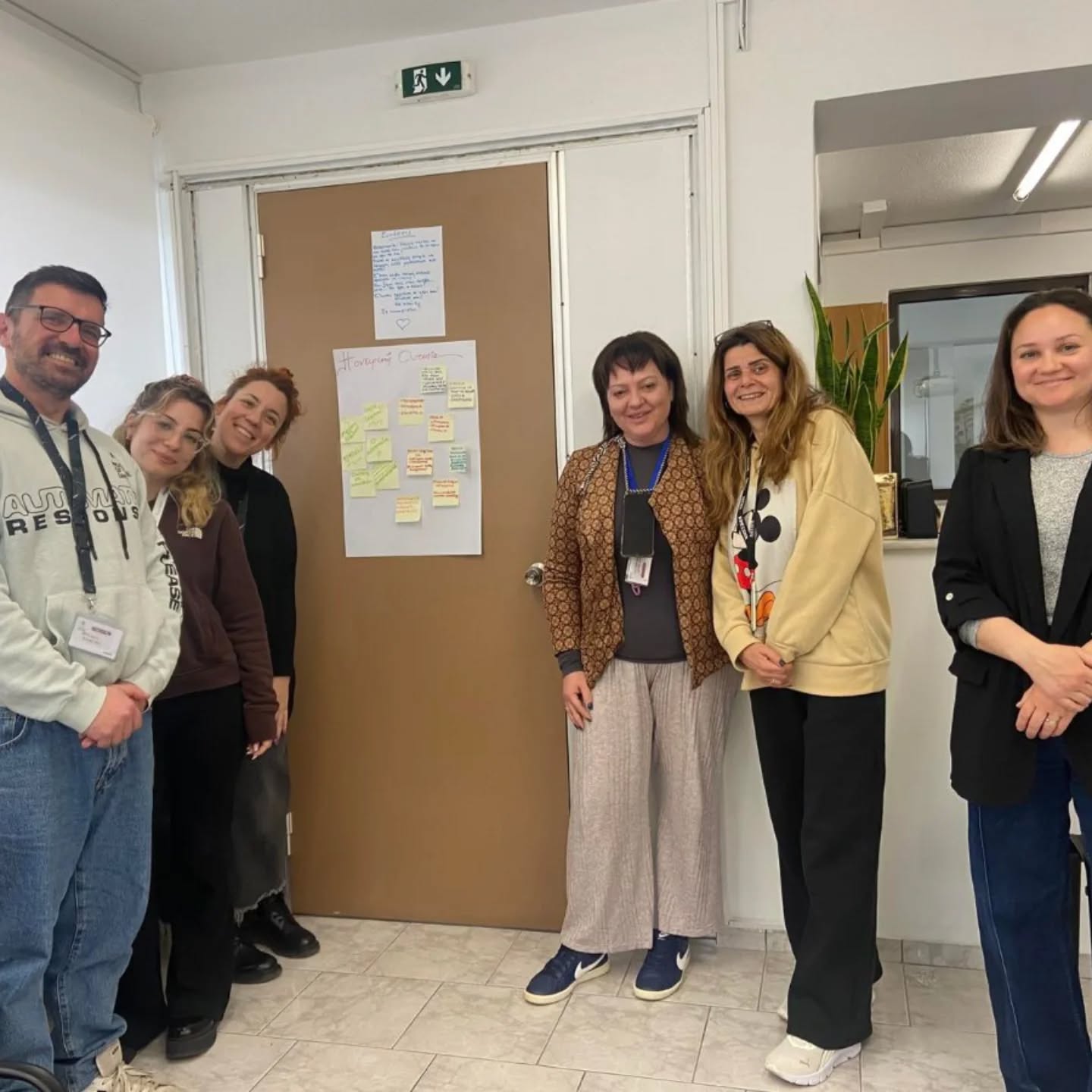Six adults stand in a hallway beside a brown door with a note-covered board and a green exit sign above. Everyone smiles for the photo, casual office attire.