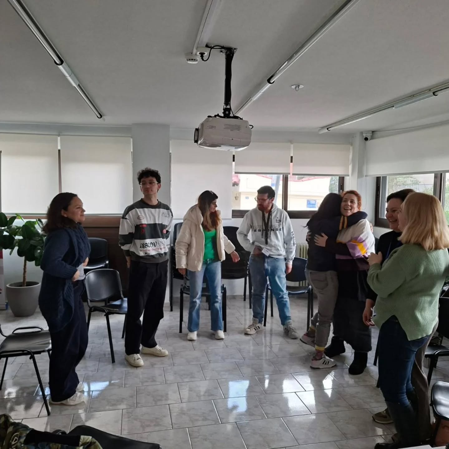 A group of people standing in a circle in a bright room, some chatting and hugging near a ceiling-mounted projector.