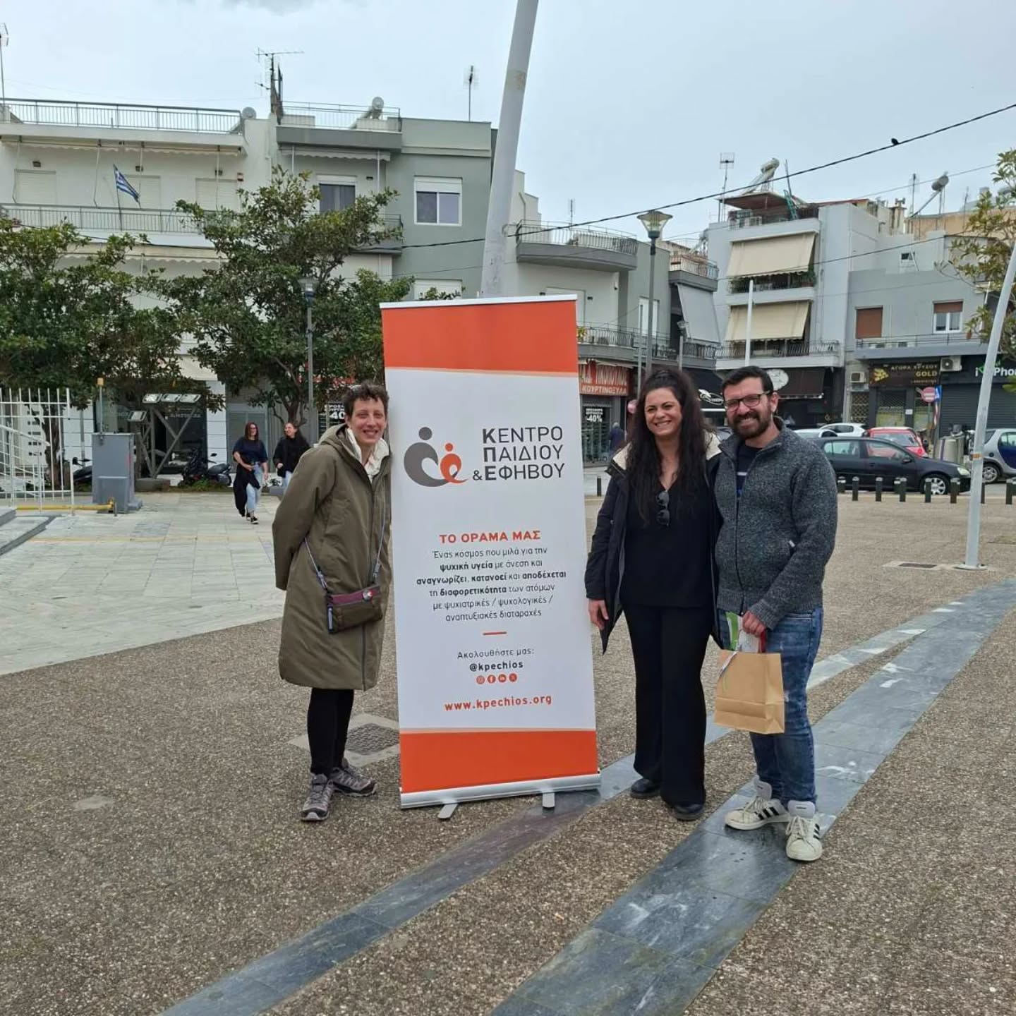 Three adults posing beside an orange-and-white banner for a children and adolescents center in a public square with buildings and trees behind them.