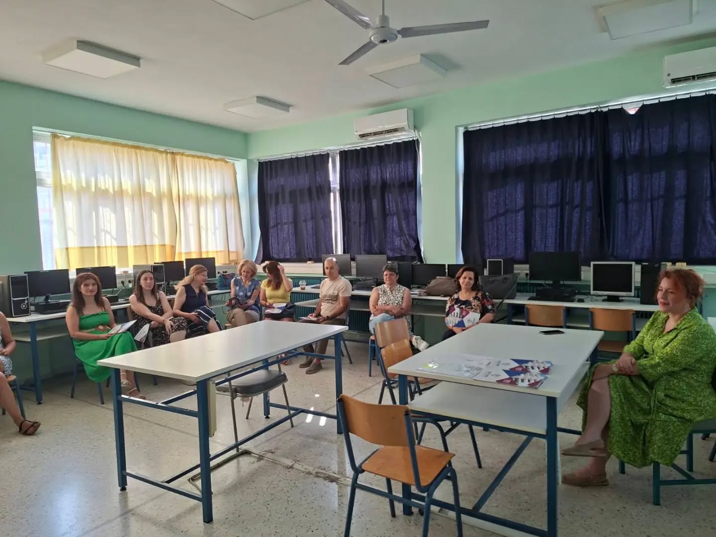 Adults seated in a computer lab arranged in a semicircle, with desktops along the back wall, listening to a session.